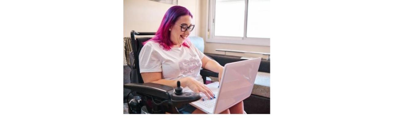 color photo of a white woman with pink hair, black glasses and a white top sitting in a wheelchair working at a computer.