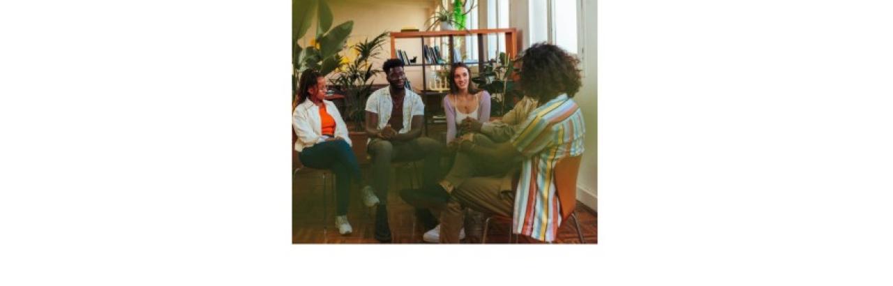 color photo of several people sitting in a circle talking. The people are Black or white and all are young.