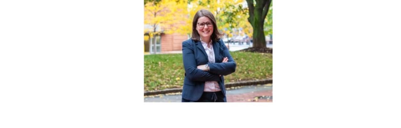 color photo of Carli Friedman, a young white woman with straight brown hair and glasses wearing a blue blazer and white shirt. She is standing with her arms crossed and is smiling. She is outside on what looks like a college campus.