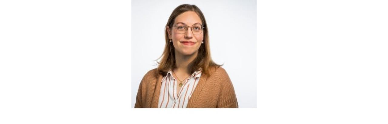 Color photo of Dalia De Santis, a young whit woman with straight brown hair and glasses wearing a tan cardigan with a red and white striped button-down short under it.
