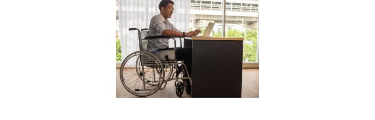 A photo of am Asian man in a wheelchair at a desk working on his laptop