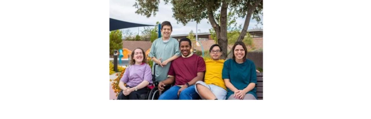five young adults sitting in a row wearing different colored solid shirts. Some have developmental disabilties