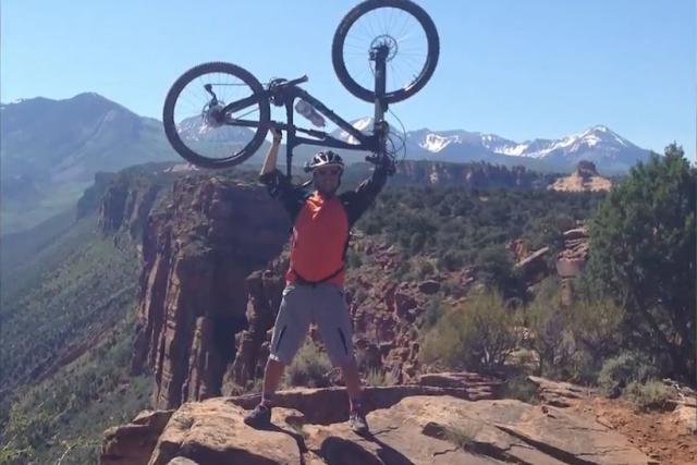 Sami on a mountain joyously holding a bike above his head