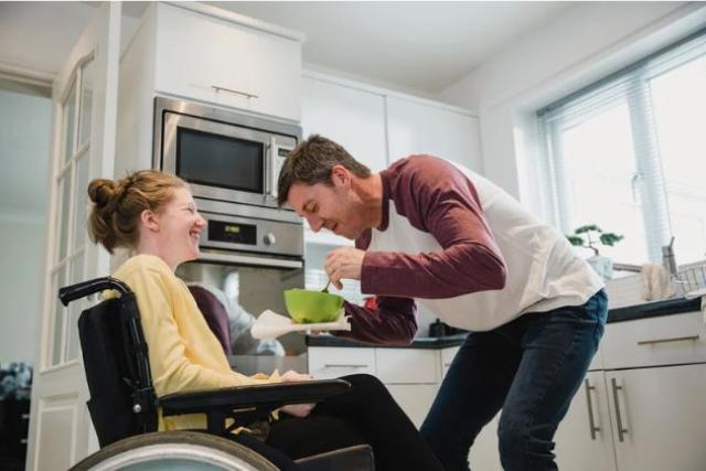 A young woman with red hair in a bun wearing a yellow sweatshrit sits in a wheelchair as a young man in jeans and a red and white shirt hold a green bowl in front of her.