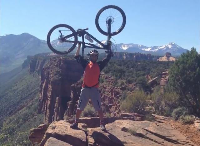 Sami on a mountain joyously holding a bike above his head
