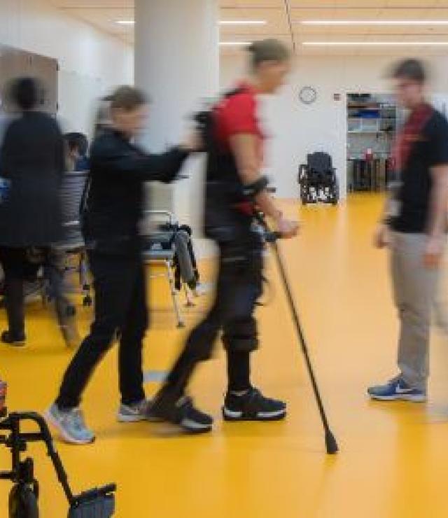a man with leg braces and crutches is walking on a yellow floor being assisted by two clinicians
