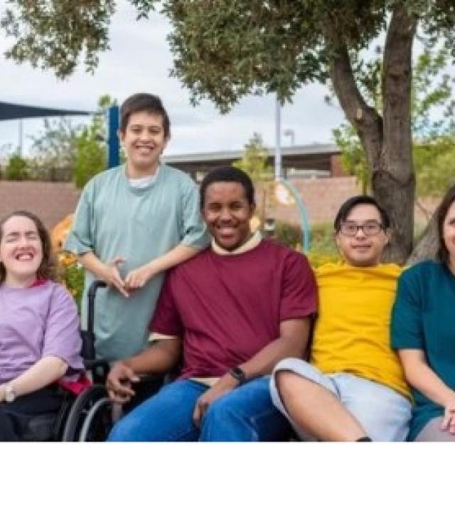 five young adults sitting in a row wearing different colored solid shirts. Some have developmental disabilties
