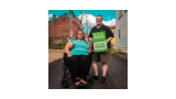 A person with a disability is sitting in a wheelchair while standing next to someone holding a green sign that reads "We are all human" in English and Spanish.