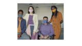 A young black man is sitting to the left of a standing white female, who is standing next to a white man sitting in a wheelchair, who is next to a standing young black woman. All of the individuals are looking into the camera and have a face mask on. 