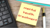 This image shows a corner of a black keyboard on the left hand side, a fountain pen on the right hand side, the head of a clipboard at the top, and in the center a post-it note that reads "Mental Health Treatment" in red ink.