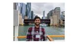 This is a picture of Dhrumil Shah. He is an Indian man who is wearing a Red flannel shirt. Behind him are a series of skyscrapers in Chicago.