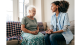 two black women sit on a couch, one older with white hair and one younger. They are smiling at each other