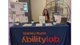 woman seatured behing a conference table with a closth that says shirley ryan abilitylab. the table has papers and pens and the woman has long brown hair and is smiling.