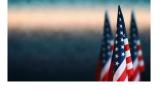 a color photo of the American flag against a blurred background of gray, white and blue sky