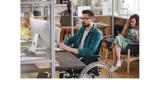 a young man with black hair and a beard is sitting in a wheelchair at a desk typing on a computer
